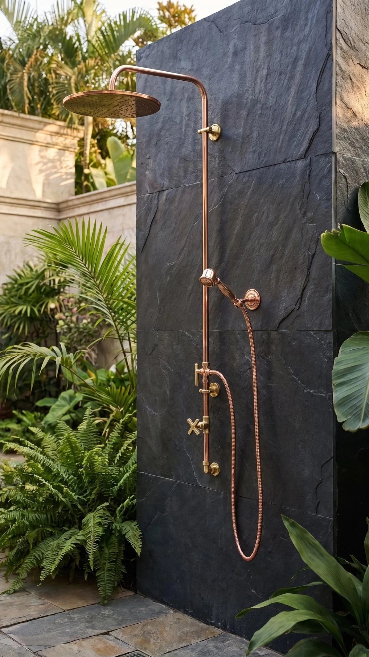 A copper outdoor shower system with a round rain shower head, adjustable wall-mounted arm, and handheld shower wand mounted on a dark slate stone wall, surrounded by lush green tropical plants.