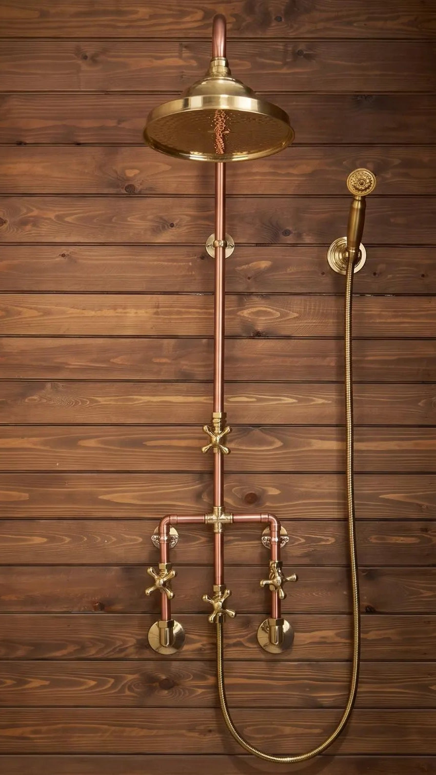 A vintage-style brass shower fixture with a large round overhead rain shower head, a vertical pipe, a hand shower, and two cross-handle valves, mounted against a rustic dark brown wood plank wall.