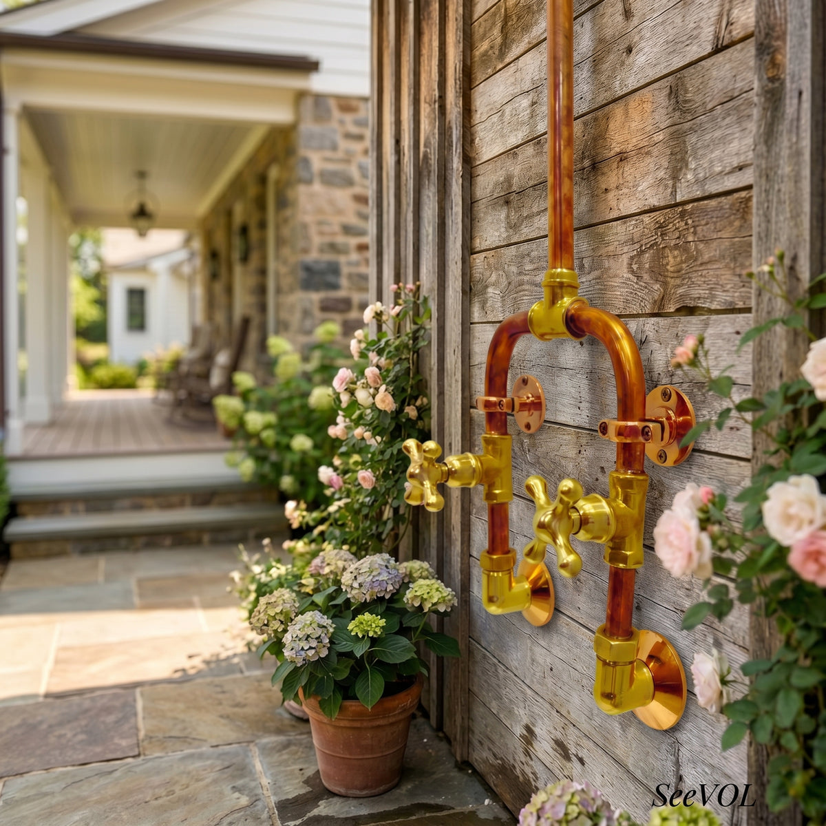 A decorative outdoor wall-mounted faucet fixture made of polished copper and gold-toned metal, mounted on a rustic wooden fence next to potted pink and white flowers. The background shows a blurred stone house porch.